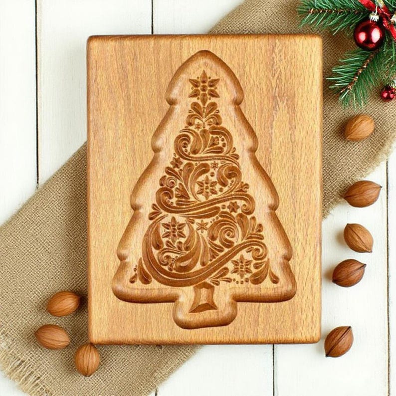 May include: A wooden Christmas tree-shaped mould with detailed floral and swirl carvings. The mould is made of light brown wood and rests on a burlap cloth. Several pecans and a sprig of evergreen with red baubles are also present.