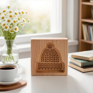 May include: A wooden block with a carved design of a winter hat. The hat design is detailed with a textured pattern. The block is light brown and sits on a white surface, with a cup of coffee and flowers in the background.