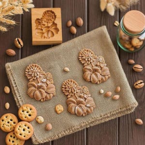 May include: A wooden cookie mold and several stamped cookies with a floral design are displayed on a tan cloth. The cookies are brown and have a detailed flower and leaf pattern. A jar of cookies and nuts are also present.