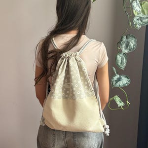 May include: A drawstring backpack featuring a sage green upper section with a white daisy pattern and a solid beige lower section. The bag includes thick, braided white rope straps and a small rectangular label with text on the front panel.