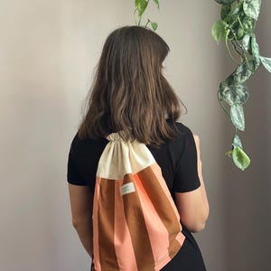 Può includere: Zaino con coulisse a righe verticali marroni e pesca, e una parte superiore color crema. La borsa è indossata sulla spalla. Una piccola etichetta bianca con testo è visibile sul davanti.