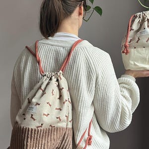 Può includere: Zaino beige con coulisse e motivo ripetuto di bassotti marroni. La sezione inferiore è in velluto a coste marrone chiaro. Lo zaino ha spallacci in corda color corallo e una piccola etichetta con la scritta "soytel tasha".