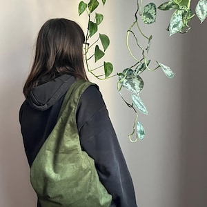 May include: An olive green shoulder bag made of a textured fabric. The bag is worn over the shoulder. The person is wearing a black hoodie and jeans. Trailing green plants are in the background.