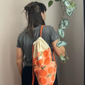 Könnte beinhalten: Ein Rucksack mit Kordelzug, der einen cremefarbenen oberen Teil und einen unteren Teil mit einem Orangenmuster auf pfirsichfarbenem Hintergrund aufweist. Die Tasche hat leuchtend orangefarbene Kordelträger und ein kleines weißes Etikett mit schwarzem Text.