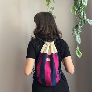 Op de afbeelding: Een rugzak met trekkoord en verticale strepen in magenta en marineblauw. Het bovenste gedeelte is crèmekleurig. De tas wordt gedragen op de rug van een persoon die een zwarte jurk draagt. Er is een klein wit label.