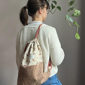 Może przedstawiać: Beżowy plecak ze sznurkiem ze wzorem brązowego jamnika na górnej części i sztruksowym dnem. Torba ma koralowe sznurki i małą etykietę. Torba jest noszona na ramieniu.