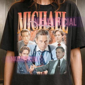 Peut inclure: T-shirt noir avec un graphisme de style vintage de Michael Douglas. Le design comprend le nom "MICHAEL DOUGLAS" dans une police dégradée orange et blanche, avec plusieurs images de l'acteur. Le t-shirt a un aspect usé et vieilli.