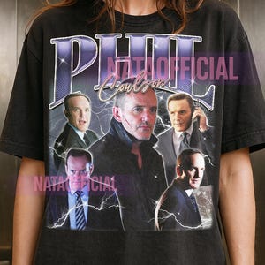 May include: A black short-sleeved t-shirt featuring a graphic print of Phil Coulson in various poses. The text PHIL is printed in large purple letters at the top, with Coulson written in script below. The design includes white lightning bolt accents.