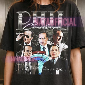 Può includere: Una t-shirt nera a maniche corte con una stampa grafica di Phil Coulson in varie pose e abiti. Il testo PHIL COULSON è stampato in un carattere argento metallizzato nella parte superiore, con accenti a forma di fulmine attorno alle immagini.