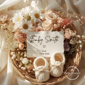 May include: A woven basket filled with white and peach-colored flowers, including roses and daisies, surrounds a card that reads "Baby Smith" with a heart and "DUE JUNE 2026." Two white knitted baby booties with ribbon bows sit in front.