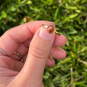 Könnte beinhalten: Ein goldener Ring mit einem runden, bernsteinfarbenen Stein. Der Ring wird vor einem verschwommenen grünen Hintergrund gehalten, der das schlichte, aber elegante Design des Rings hervorhebt. Das Ringband ist dünn und zart.