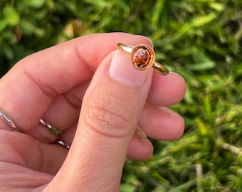 May include: A gold ring with a round, amber-colored stone. The ring is held against a blurred green background, showcasing the ring's simple yet elegant design. The ring's band is thin and delicate, highlighting the central stone.