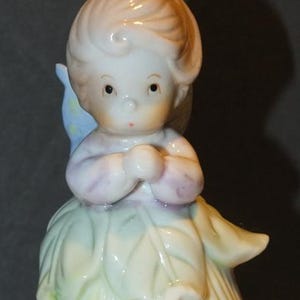 May include: A porcelain figurine of a cherubic figure with a floral dress. The figurine has a light skin tone, with a light brown hair and a pale green and yellow dress. The figurine is a trinket box with a gold band.