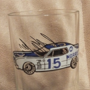 May include: A clear glass drinking glass with a blue and white illustration of a race car with the number 15 and the name "MORRIS" on the side. The car is signed by the driver.