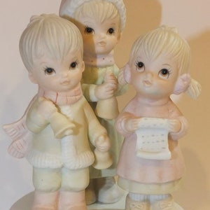 May include: A porcelain figurine of three children dressed in winter clothing, singing carols. The figurine is a musical piece with a base decorated with holly leaves and berries.