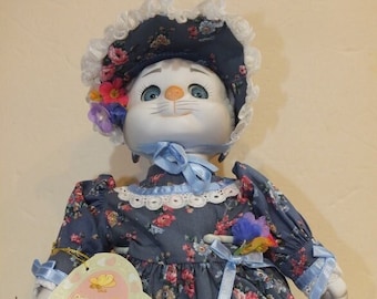 Puede incluir: Una muñeca de porcelana con cara de gato, vestida con un vestido floral azul y blanco, un sombrero de encaje blanco con cinta azul y lazos de cinta azul en sus zapatos. Está sosteniendo una tarjeta en forma de corazón con una imagen de dos gatitos.