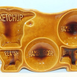 May include: A brown ceramic cow-shaped serving tray with five compartments. The compartments are labeled "Ketchup", "Hamburger", "Relish", "Hamburger", and "Mustard".