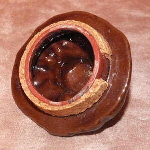 May include: A brown ceramic lid with a cork and red rim. The inside of the lid is glazed and has a textured surface.