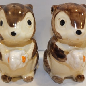May include: A pair of ceramic salt and pepper shakers shaped like squirrels. The squirrels are brown and white with black eyes and are holding a white object in their paws.