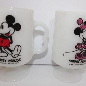 May include: Two white pedestal mugs featuring Mickey Mouse and Minnie Mouse. Mickey is in red shorts, and Minnie is in a pink polka-dot dress. Each mug has a unique handle design and the text "Mickey Mouse" and "Minnie Mouse".