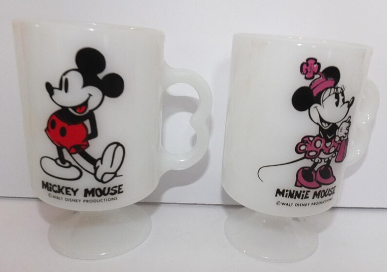 May include: Two white pedestal mugs featuring Mickey Mouse and Minnie Mouse. Mickey is in red shorts, and Minnie is in a pink polka-dot dress. Each mug has a unique handle design and the text "Mickey Mouse" and "Minnie Mouse".
