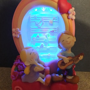 May include: A pink and orange ceramic figurine of a couple sitting on a heart-shaped couch. The man is playing a guitar and the woman is holding her hand to her cheek. The background is a blue light with musical notes.