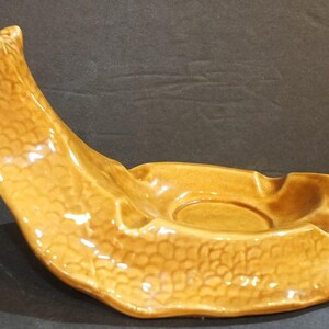 May include: A vintage, ceramic ashtray in a warm, golden-brown hue. The ashtray features a unique, curved design with a textured, scale-like pattern. It has a central circular area and two built-in rests for cigarettes.