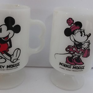 May include: Two white glass mugs featuring Mickey Mouse and Minnie Mouse. Mickey is in red shorts, and Minnie is in a polka-dot dress. Each mug has a heart-shaped handle and the text "Mickey Mouse" and "Minnie Mouse".