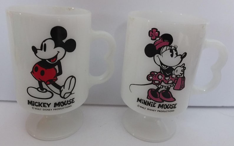 May include: Two white glass mugs featuring Mickey Mouse and Minnie Mouse. Mickey is in red shorts, and Minnie is in a polka-dot dress. Each mug has a heart-shaped handle and the text "Mickey Mouse" and "Minnie Mouse".
