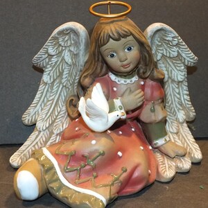 Könnte beinhalten: Eine Keramikengelfigur mit weißen Flügeln, einem Heiligenschein und einem rosafarbenen Kleid. Der Engel hält eine weiße Taube in seinen Händen. Die Figur sitzt auf einer braunen Oberfläche.