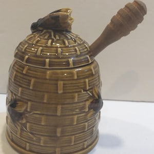 May include: A ceramic honey pot in the shape of a beehive, with a matching lid and a wooden honey dipper. The pot is brown with a woven basket design and bee accents. The honey dipper is made of light brown wood.