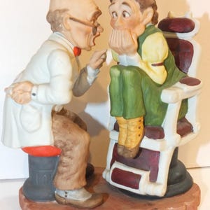 Könnte beinhalten: Eine Porzellanfigur-Gruppe, die einen Arzt in einem weißen Kittel und einen Patienten zeigt, der auf einem Friseurstuhl sitzt. Der Arzt zeigt auf den Mund des Patienten, und der Patient bedeckt seinen Mund mit seinen Händen. Die Figur ist in realistischen Farben bemalt und hat ein detailliertes Design.