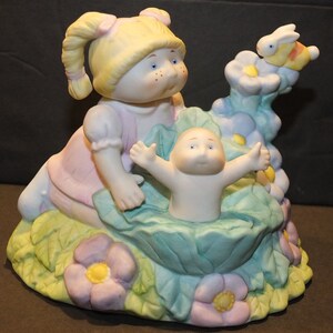May include: A porcelain figurine of two Cabbage Patch Kids dolls. The girl doll is wearing a pink dress and has blonde hair in pigtails. The boy doll is wearing a white outfit and has blue eyes. The dolls are sitting in a bed of flowers with a butterfly on a flower.