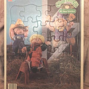 May include: A 25-piece puzzle featuring three Cabbage Patch Kids dolls. The dolls are dressed in cowboy outfits and are standing in a field of hay. The puzzle is designed for ages 3 to 5.