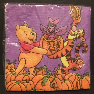 May include: A package of Halloween-themed paper napkins featuring Winnie the Pooh, Piglet, and Tigger. The characters are holding a carved pumpkin. The background is purple, and there are orange pumpkins at the bottom.