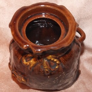 May include: A brown ceramic jar shaped like a sack with a gold ribbon and a brown lid. The jar has a textured surface and a gold inscription that reads "Merry Christmas".