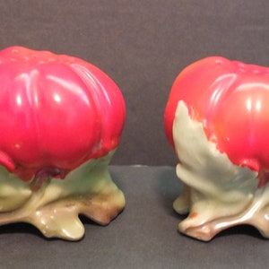 May include: A pair of red ceramic tomato salt and pepper shakers with green leaves and brown stems.