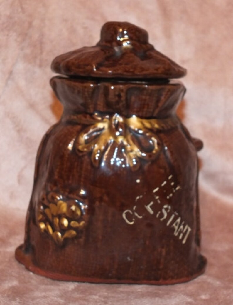 May include: A brown ceramic coffee jar shaped like a sack with a lid. The jar has gold accents and the text "Coffee" and "Constant" written on it.