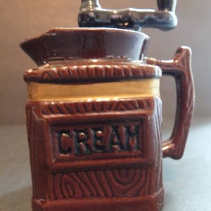 K&ouml;nnte beinhalten: Ein brauner Keramik-Sahnegie&szlig;er mit Holzmaserung und schwarzem Griff. Der Sahnegie&szlig;er hat oben eine Handkurbel und das Wort "CREAM" steht auf der Vorderseite.