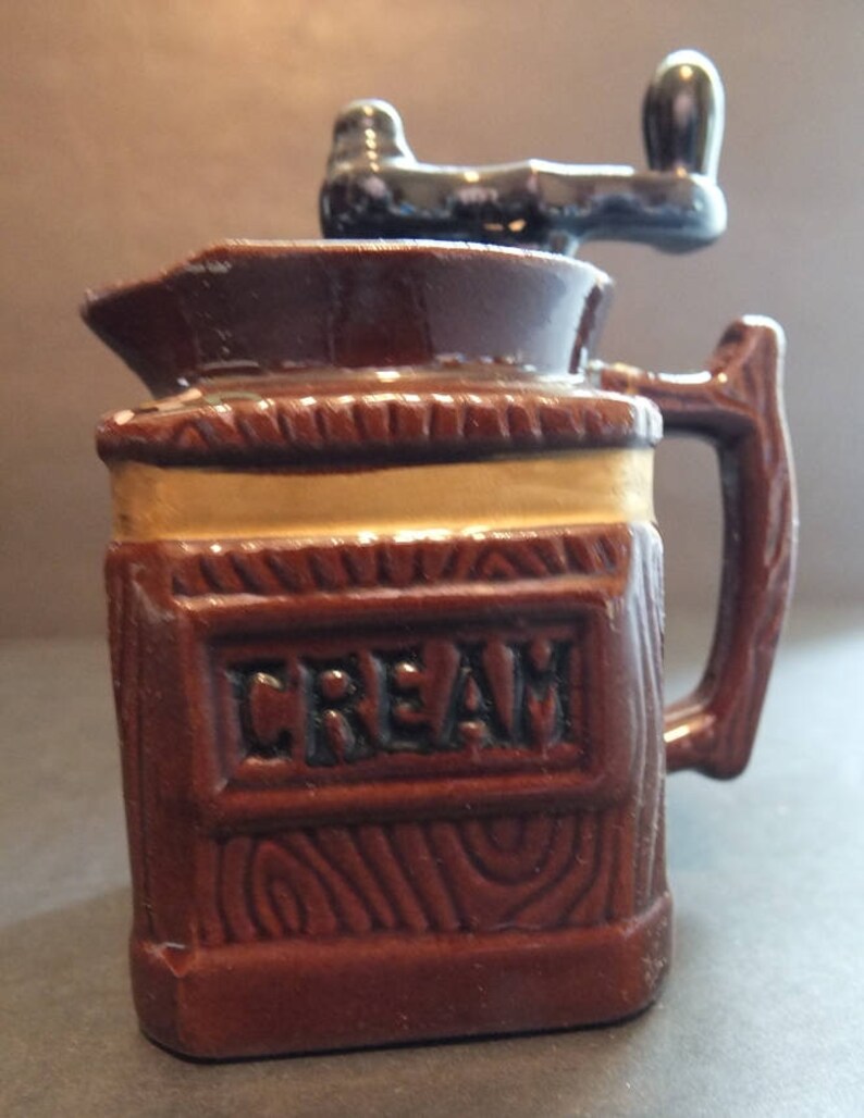K&ouml;nnte beinhalten: Ein brauner Keramik-Sahnegie&szlig;er mit Holzmaserung und schwarzem Griff. Der Sahnegie&szlig;er hat oben eine Handkurbel und das Wort "CREAM" steht auf der Vorderseite.
