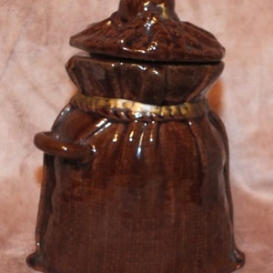 May include: A brown ceramic jar shaped like a sack with a lid. The jar has a gold rope detail around the top and a handle on the side.