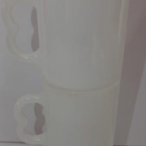 May include: Two stacked, white, vintage-style mugs. Each mug has a heart-shaped handle and a tall, cylindrical body. The mugs are made of a translucent material, and the bottom mug sits on a small, round base.