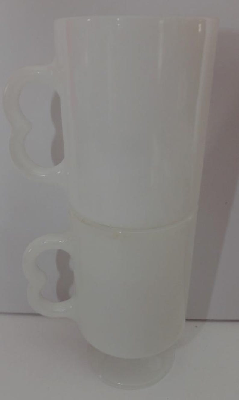 May include: Two stacked, white, vintage-style mugs. Each mug has a heart-shaped handle and a tall, cylindrical body. The mugs are made of a translucent material, and the bottom mug sits on a small, round base.