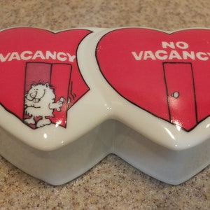 Könnte beinhalten: Eine herzförmige Keramik-Schmuckschatulle. Die weiße Basis trägt zwei rote herzförmige Deckel. Einer trägt die Aufschrift "VACANCY" mit einer Cartoonfigur, der andere "NO VACANCY".