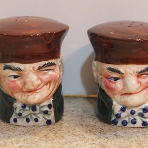 May include: A pair of ceramic salt and pepper shakers shaped like the heads of men wearing brown hats and green jackets. The men have white faces with rosy cheeks and are winking.