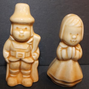 Könnte beinhalten: Zwei Keramikfiguren in hellbrauner Glasur. Eine ist ein Soldat mit Hut und Umhang, die andere ein Mädchen mit gefalteten Händen. Die Figuren haben ein Vintage-Aussehen, mit einfachen Gesichtszügen und detaillierter Kleidung.