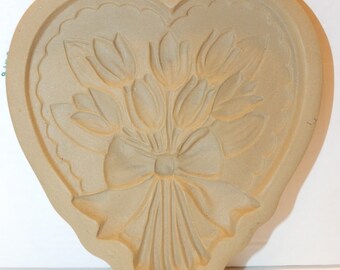 May include: A heart-shaped cookie cutter with a design of a bouquet of tulips tied with a bow. The cutter is made of beige ceramic and has a green ribbon attached.