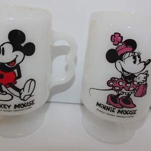 May include: Two white glass mugs featuring Mickey Mouse and Minnie Mouse. Mickey is in red shorts, and Minnie wears a pink dress and bow. Each mug has a curved handle and the text "Mickey Mouse" and "Minnie Mouse".