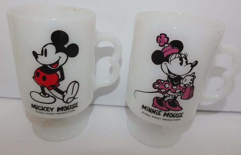 May include: Two white glass mugs featuring Mickey Mouse and Minnie Mouse. Mickey is in red shorts, and Minnie wears a pink dress and bow. Each mug has a curved handle and the text "Mickey Mouse" and "Minnie Mouse".