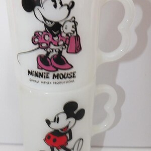 May include: Two white glass mugs featuring Minnie Mouse and Mickey Mouse. Minnie is depicted in a pink dress and bow, while Mickey wears red shorts. The mugs have heart-shaped handles and the text "Minnie Mouse" and "Mickey Mouse".